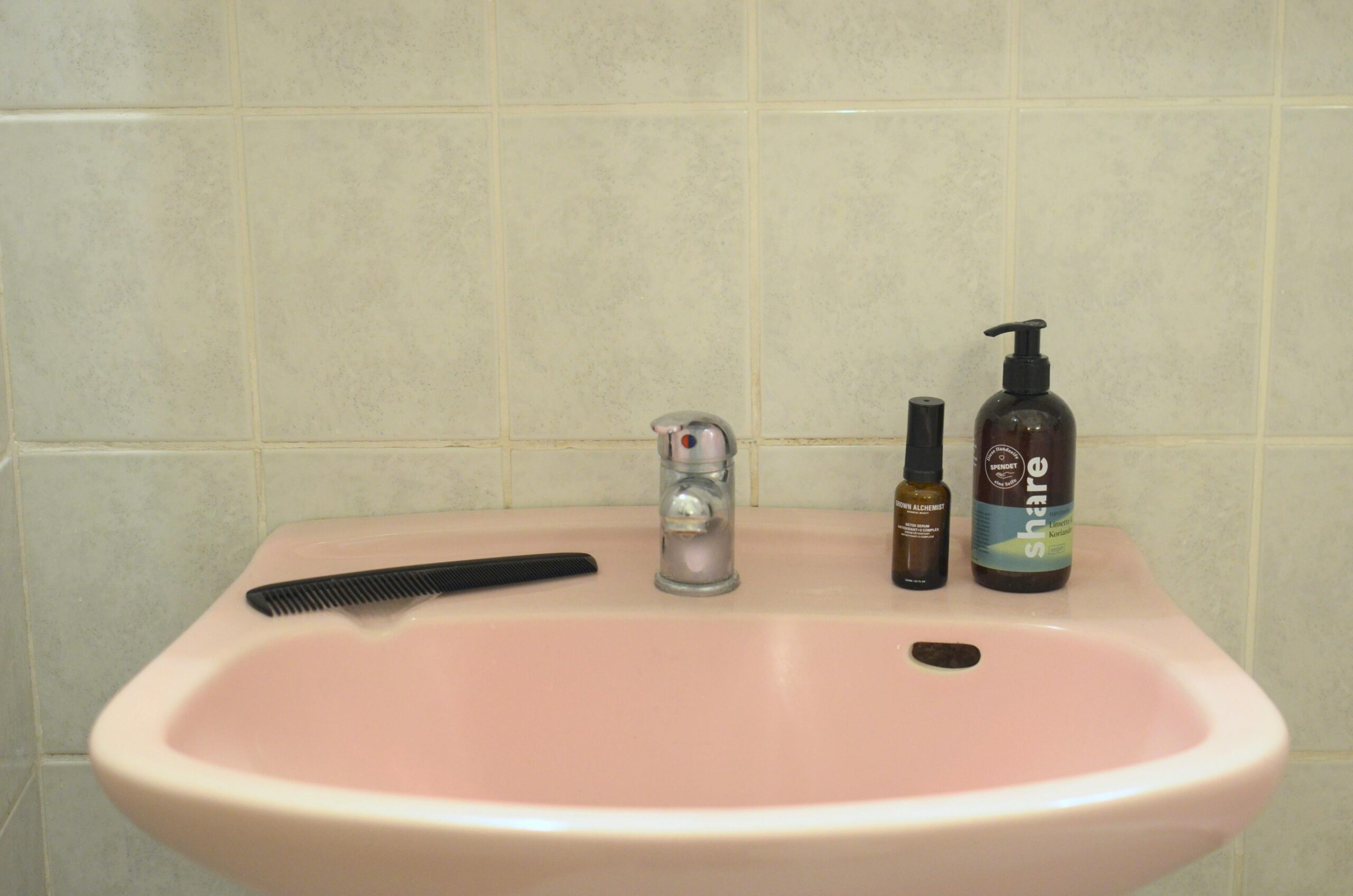 A pink ceramic washbasin with hygiene products and a comb on a tiled wall.